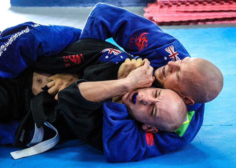 A BJJ athlete takes the back of another and proceeds to set up a choke.
